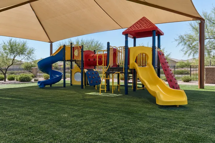 Artificial turf under a shaded commercial playground structure at a community park, safety-colored equipment without text