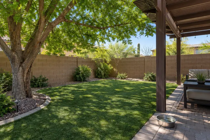Backyard corner with artificial turf under the shade of a mature tree and a pergola, a water bowl on the patio edge